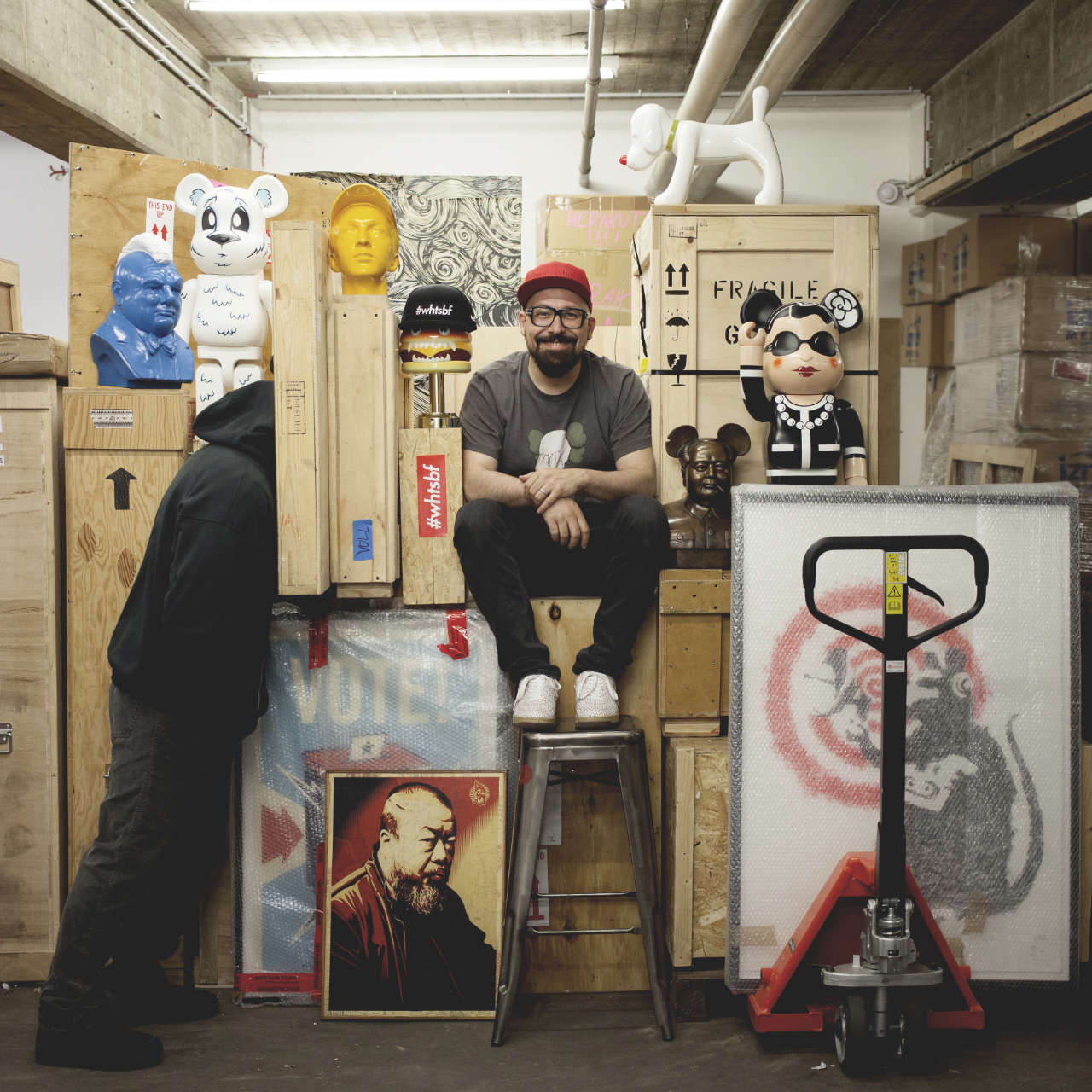 Das Foto zeigt Selim Varol, der auf Kisten in einer Art Lager sitzt und grinst. Er trägt eine rote Kappe und ein dunkelgraues T-Shirt mit einem Motiv des Künstlers KAWS. Um ihn herum sind verpackte Kunstwerke – Figuren und Bilder – zu erkennen.